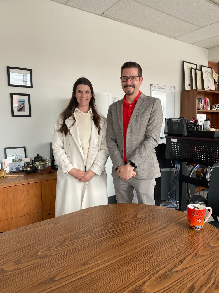 Emma stands beside Dr. Thomas Piggott, in front of a board room table with office accoutrements in the background, and a beautiful red/orange coffee mug in the foreground.
