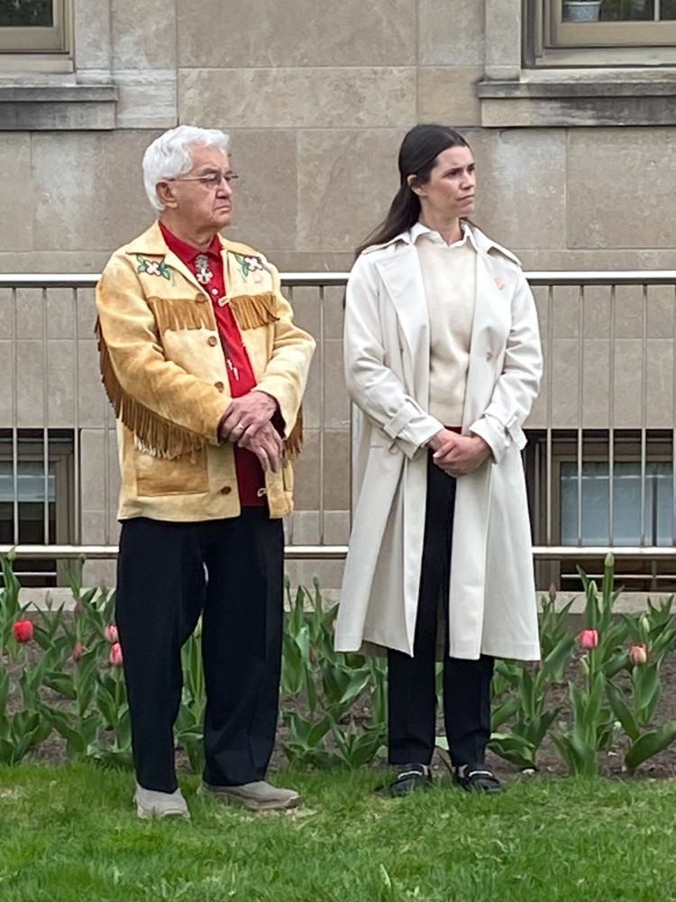 Emma stands beside Curve Lake First Nation Chief Keith Knott on the lawn at Peterborough City Hall, with spring tulips in the background.