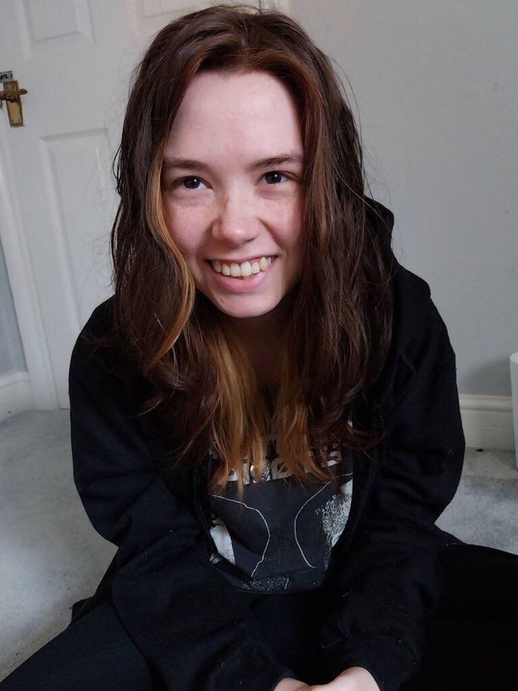 photo of myself smiling sat cross legged indoors on a carpeted floor with my hands in my lap. im a white, feminine looking young adult, with long brown hair with some blonde streaks that is a little wet in the photo. im wearing a black hoodie, grey twenty one pilots merch and black jeans