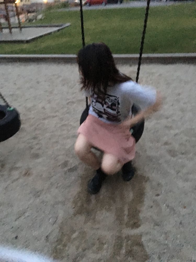 blurry picture of a figure with dark hair in t shirt and comfy skirt in a tyre swing turned away from the camera, in slightly low lighting but still looks like daylight