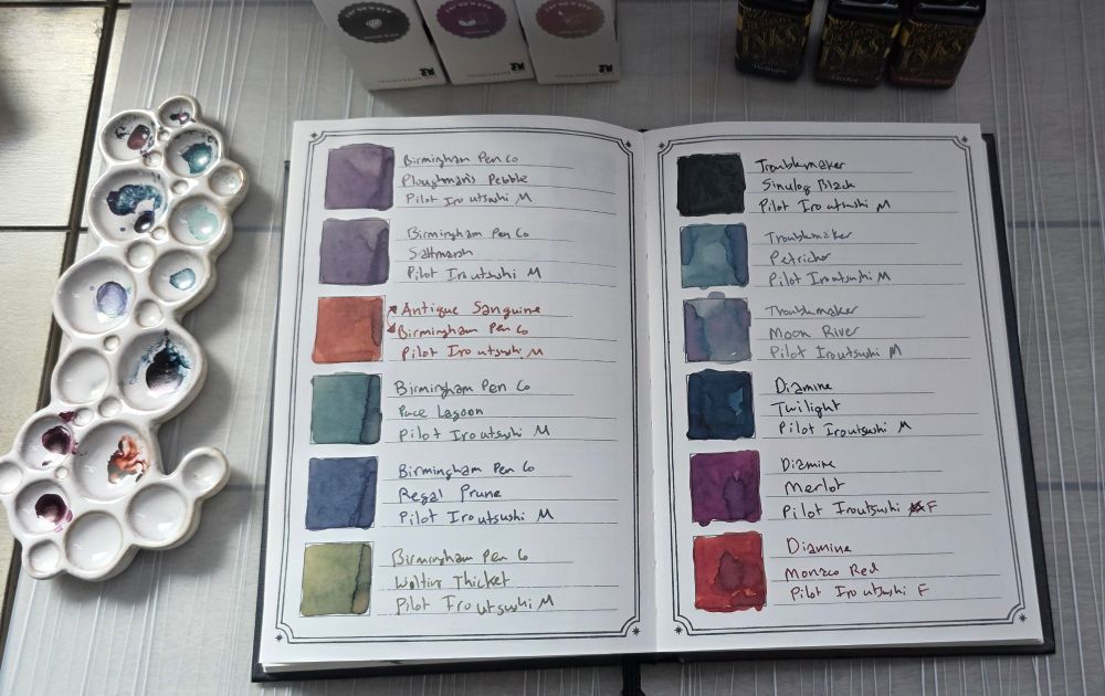 A ceramic paint palette that looks like a cluster of bubbles in varying sizes next to the Dominant Industry Ink Archiving Book, A Log of Elixir, open to a page showing ink swatches for six Birmingham Pen Co inks and these inks:

Troublemaker Sinulog Black, a dark greenish black
Troublemaker Petrichor, a chromashading blue, green, purple
Troublemaker Moon River, a gray, teal, and pink
Diamine Twilight, a dark blue
Diamine Merlot, a pinkish purple
Diamine Monaco Red, a warm muted