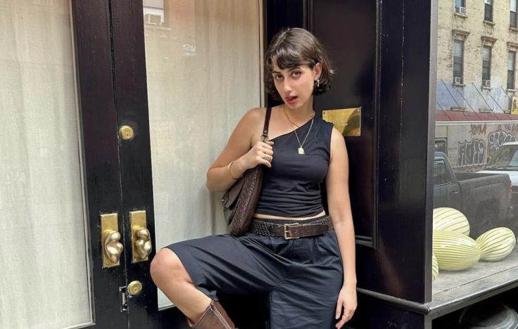 Zohran Mamdani's wife, Rama Duwaji, who is a professional artist. She has short brown hair, an earring on her left ear, and is sticking her tongue out a little. She's wearing a black top with only one shoulder and black shorts, and is holding her right leg up. She also appears to be wearing brown boots but the picture is cropped so that you can only see the top of one of them.