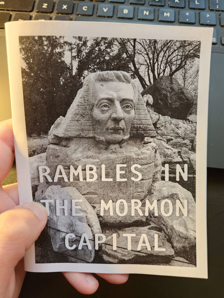 A hand holding a zine. The cover of the zine is in black and white and depicts a sphinx with the face of Joseph Smith. The cover reads "Rambles in the Mormon capital."