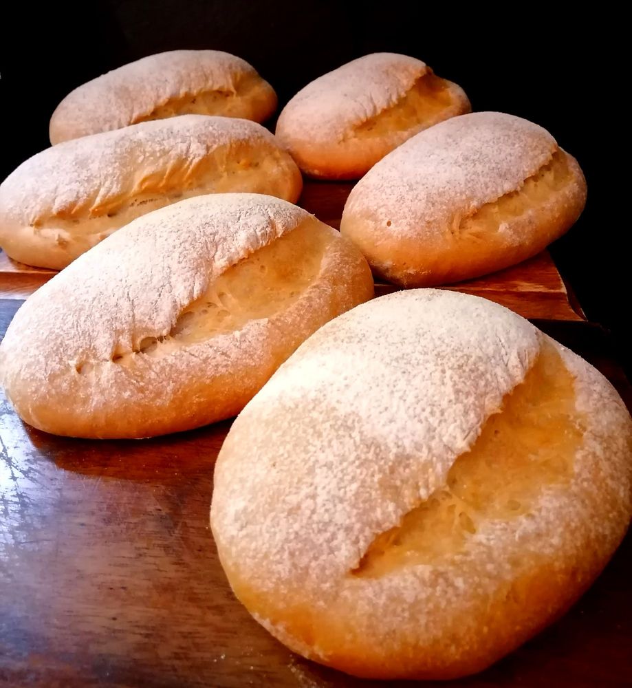 Six golden brown, flour dusted, mini loaves of bread on wooden chopping boards. 