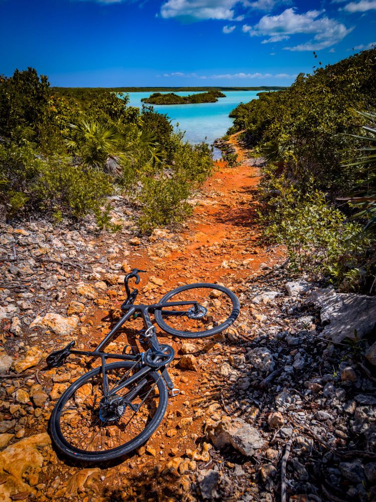 Photo of a black Scott addict gravel bike lying on the ground on a rocky red dirt trail. The trail continues towards a body of bright blue water, and then bends to the right and disappears in some green brush. In the distance, we can see small islands in the body of water. The sky above is bright blue with just a few clouds in it.
