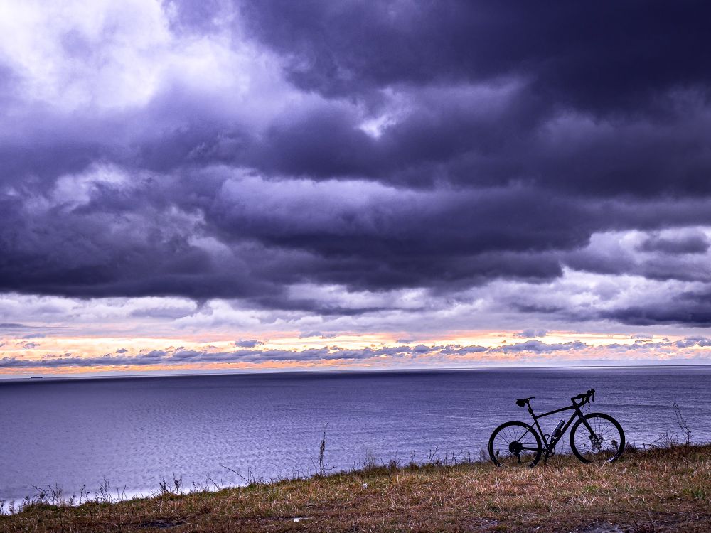 Photo of a black specialized diverge gravel bike standing on a grassy hill that is overlooking a large lake. The sky above is filled with dramatic clouds that have a purple hue. The sky is very close to the horizon is glowing bright orange. The water is also tinted purple.