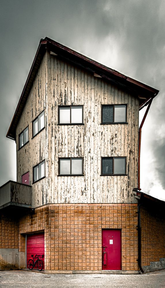 Photo of a black specialized, diverge, gravel bike standing next to the red garage door of a three-story building. The first story of the building is finished square red bricks. The top two stories are finished with yellow painted siding that is heavily worn and chipped. It’s an unusually shaped building that has three sides that we can see and a pointed roof on top of the third story. there is also a red door on the front of the building. The second and third story have small windows. The sky above is cloudy and gray.