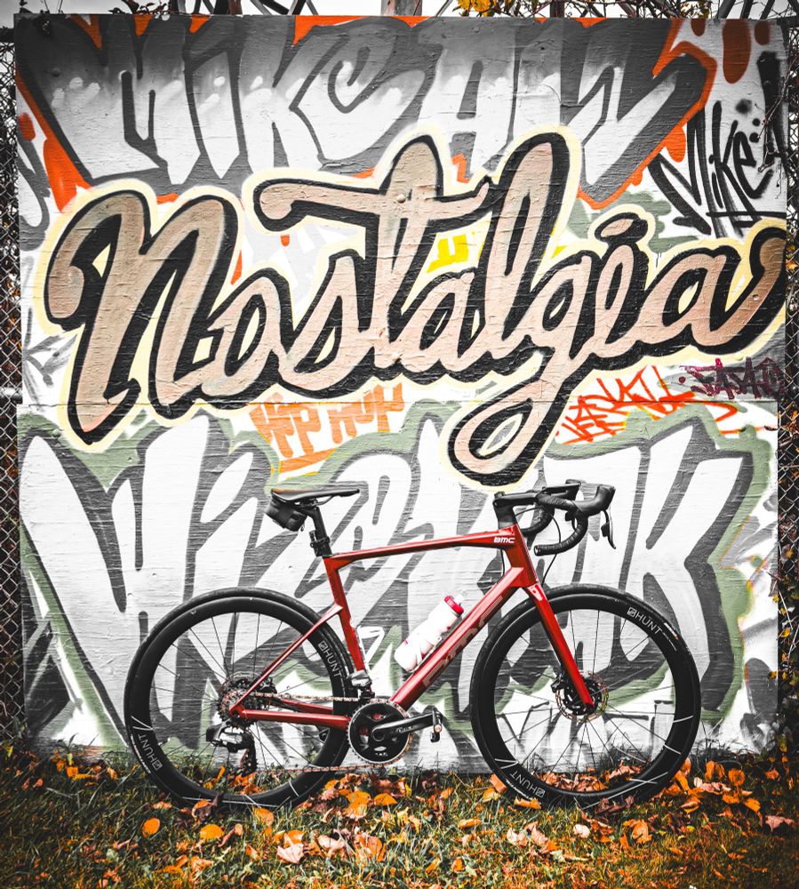 Photo of a red BMC roadmachine road bike standing next to a wall that is filled with graffiti style art. The graffiti is spelling all sorts of names and words, but prominently in the middle is the word nostalgia in cursive, gold letters. The ground beneath the bike is grassy with a light dusting of orange autumn leaves