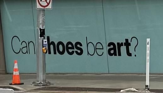 photo of the bata shoe museum in toronto. it's a glass wall with the words "can shoes be art?" on it, but the S in shoes is obscured by a metal service pole.