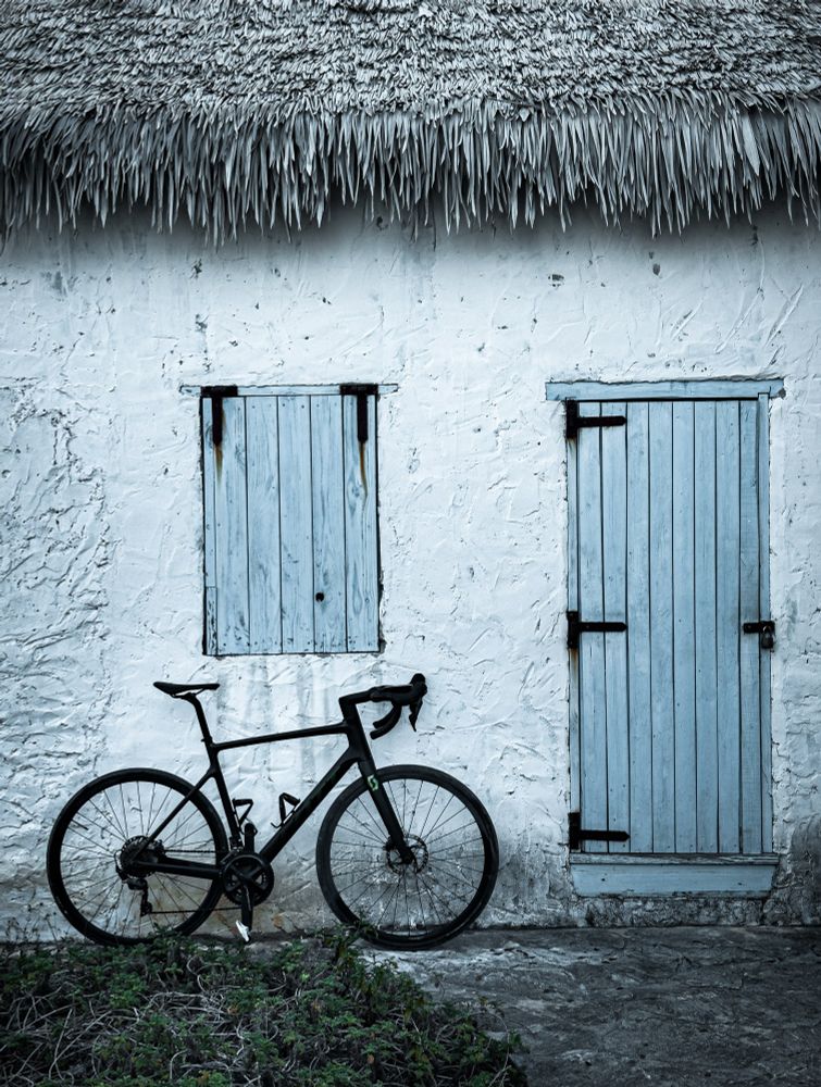 photo of a black scott addict gravel bike standing next to the wall of a building. the wall is white and textured, with a blue wooden door and a blue wooden window on it. at the top of the wall is a roof covered in grass 