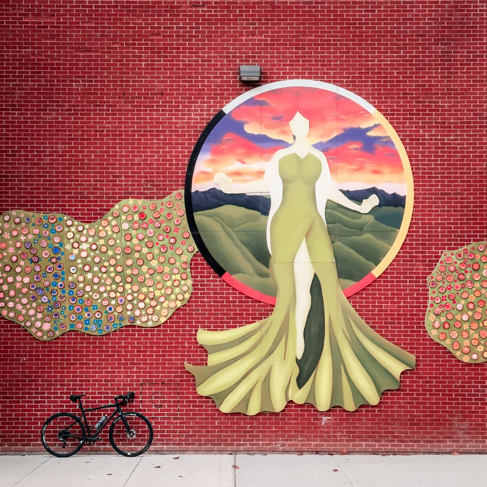 Photo of a black specialized diverge gravel bike standing up against a red brick wall that has a large piece of public art on it. The art depicts a white woman in a flowing green dress standing in front of a circle that frames a sunrise over rolling Green Hills. The frame of the circle has white yellow, red, and black colours on it. on either side of the circle or patches of green with small slices of tree branches filling them.