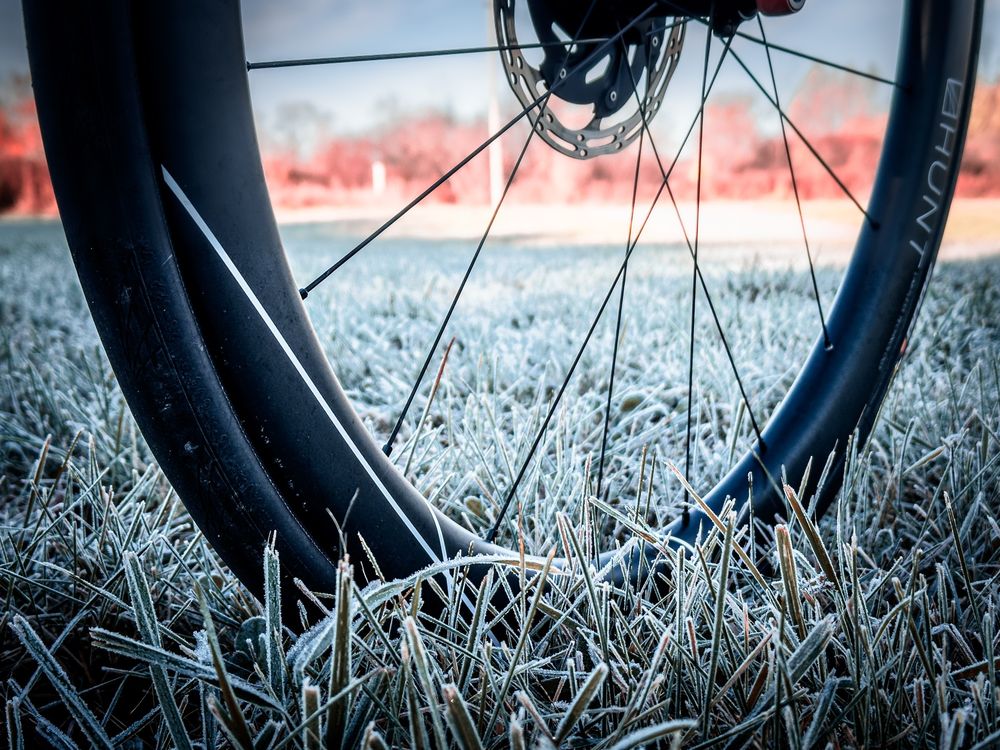 Photo of a black road bike wheel standing in uncut grass that has a blanket of frost over it. through the wheel, we can see a line of trees in the distance that is blurred and showing autumn colors. 