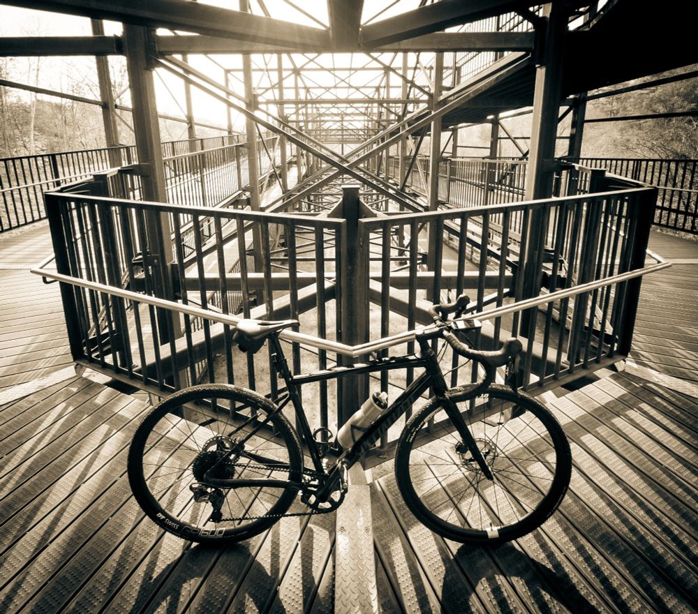 monochrome photo of a black specialized diverge gravel bike standing at the corner of a switchback metal walkway. The sun is shining through the intricate metal structure towards us so the bikes shadow is casting downwards. 