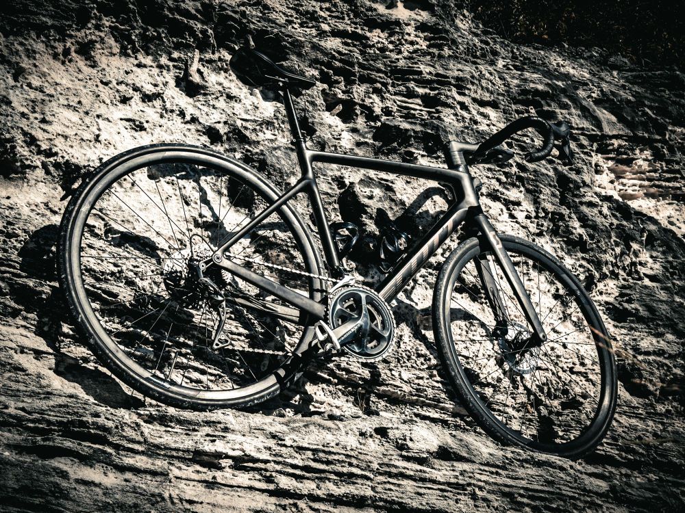 monochrome photo of a black scott addict gravel bike leaning against a rocky wall. the photo is taken from below and behind the bike, looking up at it