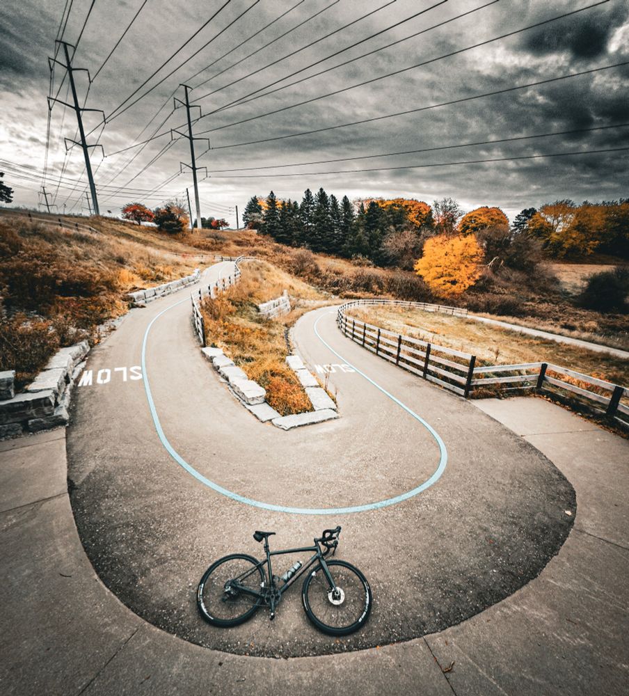 Photo of a black specialized diverge gravel bike lying on the ground in a hairpin turn on a trail that is switch backing down a hill. There are a variety of trees on the hill of the head, showing autumn colors. The grass in between the switchback is Long and Brown. The sky above is gray and showing dramatic clouds. At the top of the hill, there are electricity transmission towers, whose power lines are running straight overhead. The trail has a blue line running down the centre of it and the word “slow“ stencilled every so often