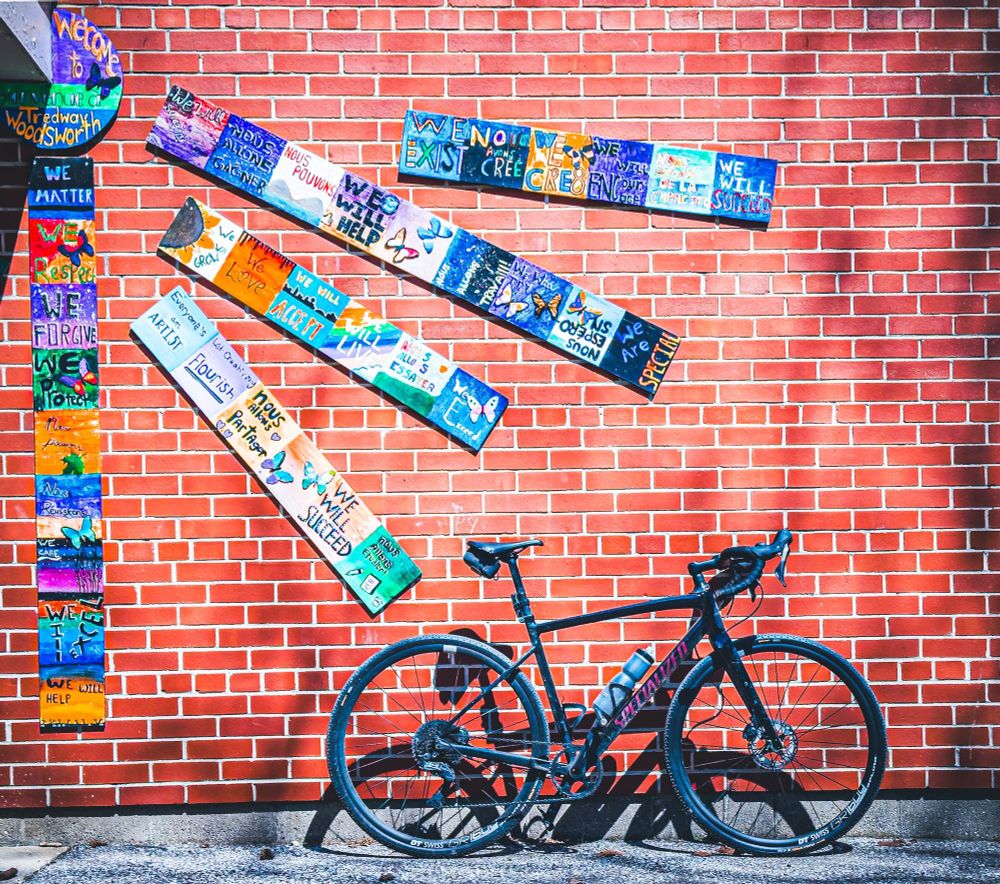 Photo of a black specialized diverge gravel bike standing next to a red brick wall. On the wall are some colourful children’s art arranged in long strips that appear to be emanating from a circle in the top left corner of the photo. 