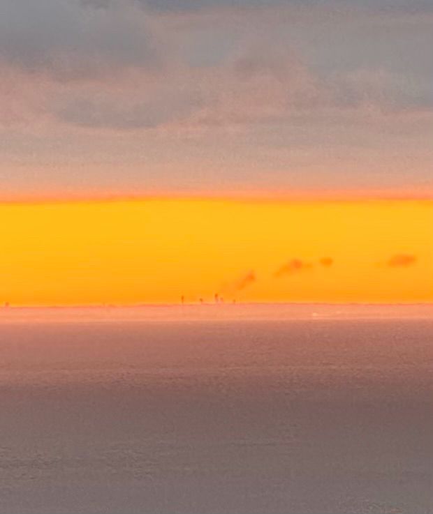 A band of bright orange sky sandwiched between medium grey sky and dark grey Lake Ontario. The towers of Niagara Falls Ontario are visible as tiny dark points at the bottom of and backlit by the orange band. 