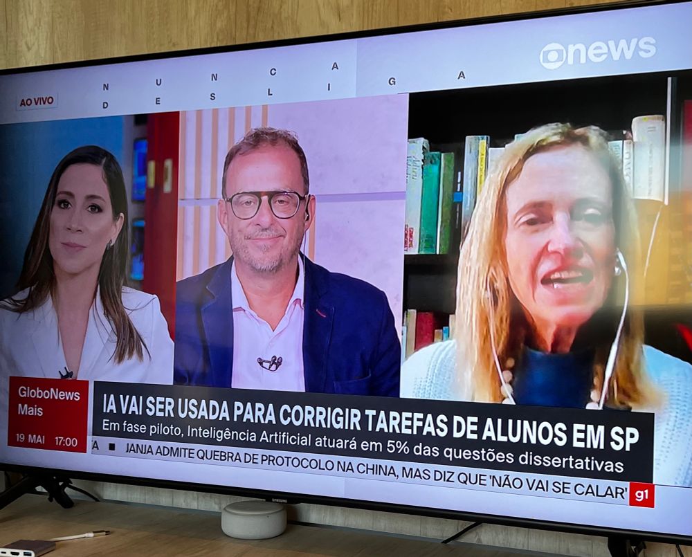 Captura de tela do canal GloboNews mostrando uma transmissão ao vivo com três pessoas em vídeo: uma apresentadora à esquerda, um homem no centro com óculos e blazer azul, e uma mulher à direita com fones de ouvido e livros ao fundo. Na parte inferior da tela, há uma manchete que diz: “IA VAI SER USADA PARA CORRIGIR TAREFAS DE ALUNOS EM SP – Em fase piloto, Inteligência Artificial atuará em 5% das questões dissertativas”. No canto inferior esquerdo, há a marca “GloboNews Mais” com a data 19 de maio às 17h00. Uma tarja de notícias rolando abaixo diz: “JANJA ADMITE QUEBRA DE PROTOCOLO NA CHINA, MAS DIZ QUE ‘NÃO VAI SE CALAR’”.