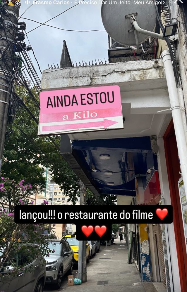 mostra uma placa de restaurante com a frase “AINDA ESTOU a Kilo” em destaque, seguida de uma seta indicando a direção. A legenda adicionada à foto brinca: “lançou!!! o restaurante do filme ❤️❤️❤️”

Além disso, a música que aparece no topo é “E Preciso Dar Um Jeito, Meu Amigo!”, de Erasmo Carlos, o que adiciona um tom dramático ou nostálgico à cena, reforçando o humor da legenda.