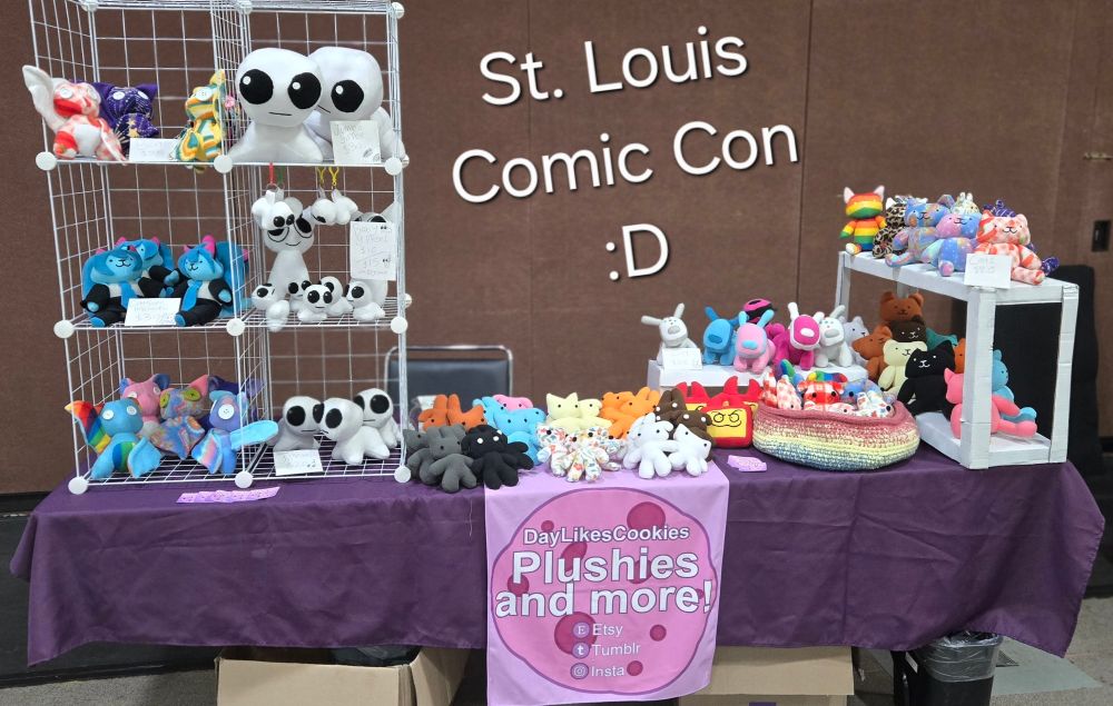 A photograph of an artist's table at a convention. It is filled with handmade plushies, including cats, bats, Hatsune Miku, iDogs, and other designs.