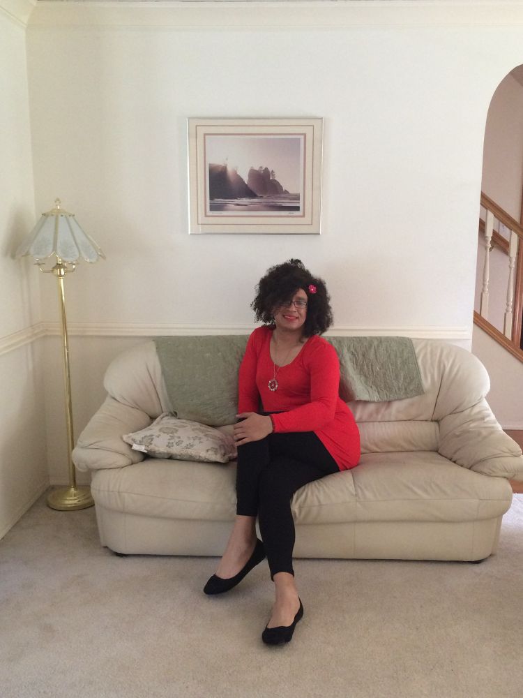 June 16, 2016 — Bianca poses on a beige couch with left leg crossed over right. I’m smiling and wearing a red long-sleeve tunic with black leggings and black flats, with a small red flower in my hair and a circular pendant making the peace sign, it has blue and pink stones on it. Notable because this was the first time I presented as my real self to family, who were already accepting but had previously done so from afar