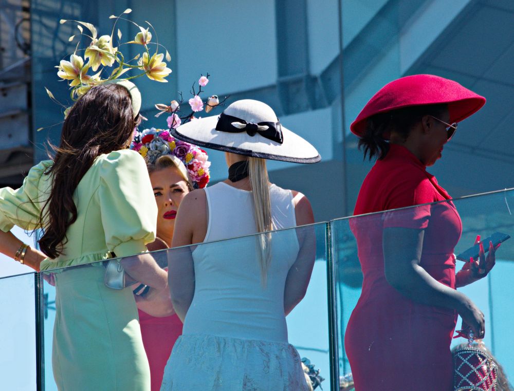 A picture of the ladies and their stunning hats at the Dundalk Races. There is a yellow flowered hat, a multi-coloured flowered hat, a wide brimmed white and black ribboned hat and a scarlet colour saucer shaped hat. 
