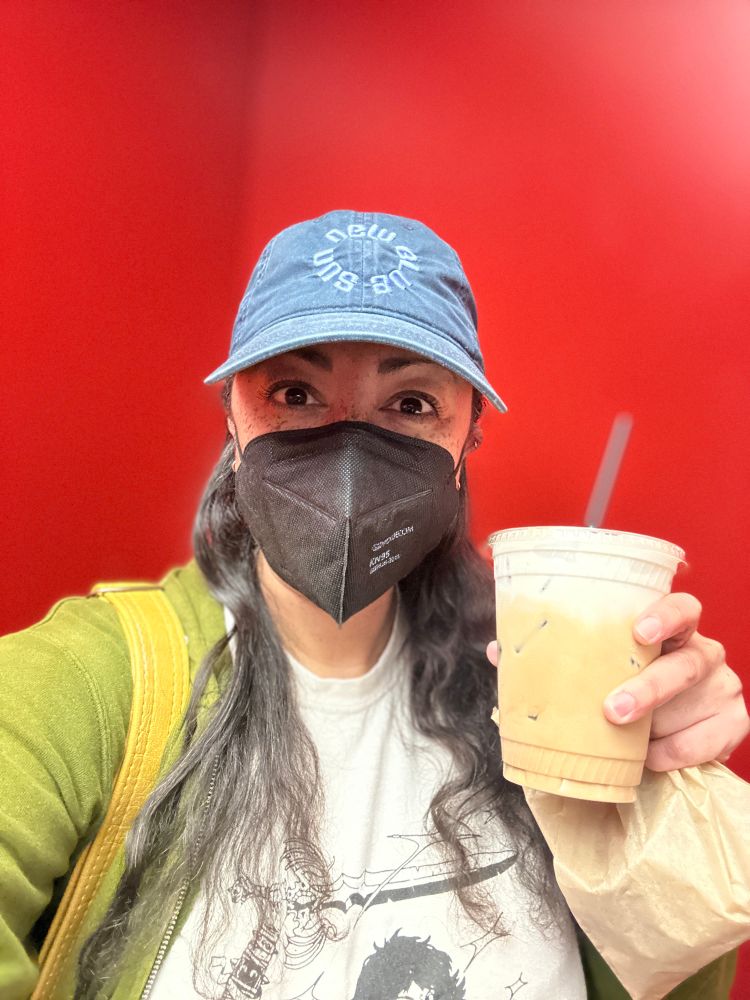 A multiracial woman wearing a black kn95, holding a coffee and pastry in a red elevator 