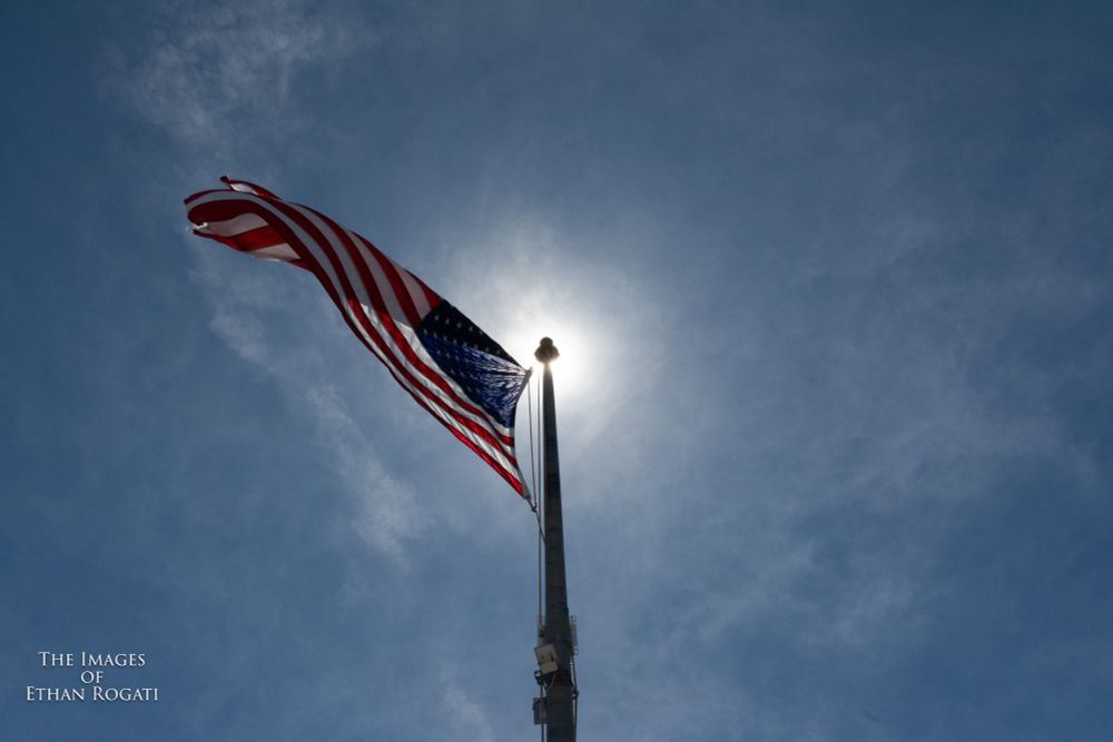 A photograph of the mid-day sun shining above an American flag, with the perspective putting the sunlight behind the top of the flag pole