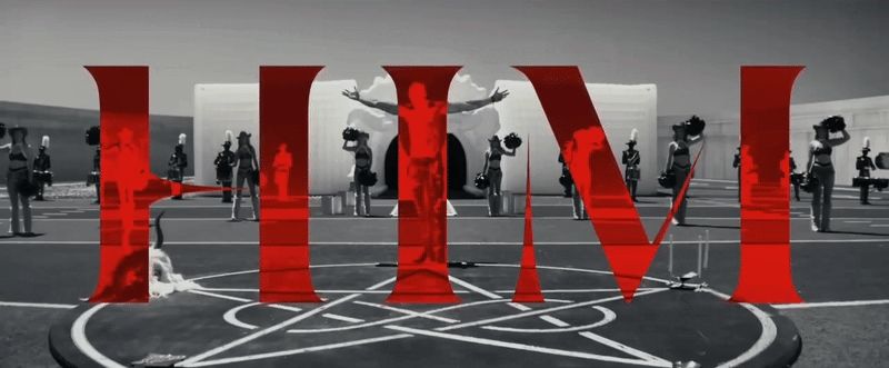 Monochromatic still from the 2025 movie Him of a man, surrounded by cheerleaders, standing in the center of the frame with his head bowed and arms outstretched with the word "HIM" across the frame in red text