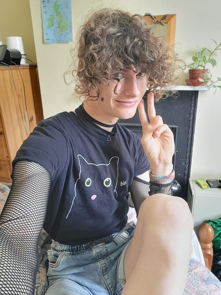 A selfie of me in a "blep" cat t-shirt with fishnet arms, in a black choker with a triangle necklace, and light blue denim shorts.