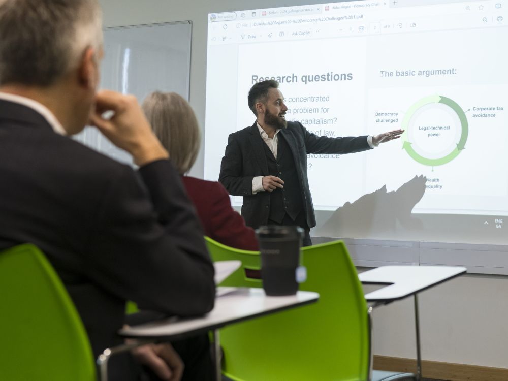 A man presenting to an audience, gesturing as he explains his topic to the engaged group of listeners.