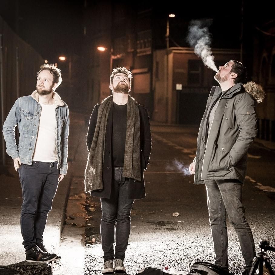A photo of IMLÉ, three white men with dark hair and beards standing on a backstreet at night, illuminated by a streetlamp. One of them is holding a cigarette, and blowing smoke into the air. (I think their line-up may have changed since this photo, and Róisín may essentially be a permanent member of the group now.)