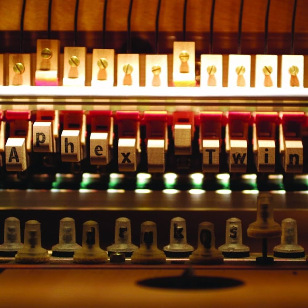 The album art for drukQs by Aphex Twin. A close-up colour photo of the innards of a 1998 Yamaha C1 Disklavier (a computer-interfacing prepared piano) used on drukQs; the words Aphex Twin are written on the damper underlevers, and drukQs on the solenoids.