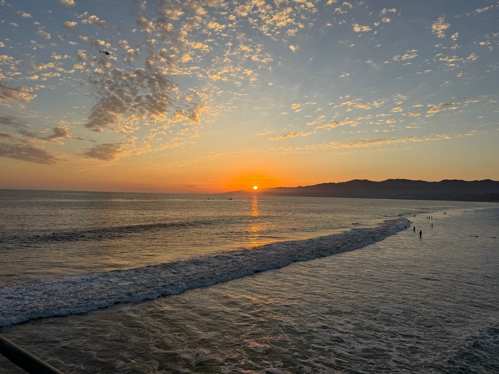 Photo of the Pacific Ocean from Santa Monica Pier, facing northwest toward the setting sun. The bottom of the sun is below mountains visible in the background.