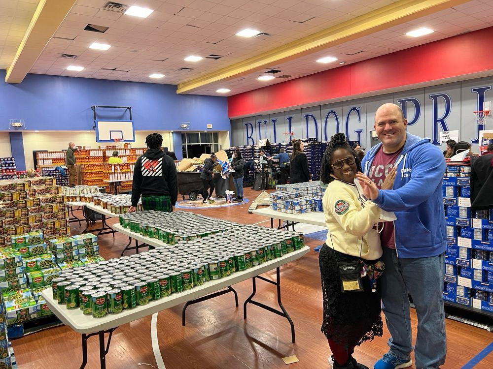 Photo of Turquoise and Greg posing for a photo in front of the gym. Behind them are several volunteers, tables full of canned vegetables and canned meats. Hundreds of boxes of cereal can be seen in the background, along with dozens of racks of bread.