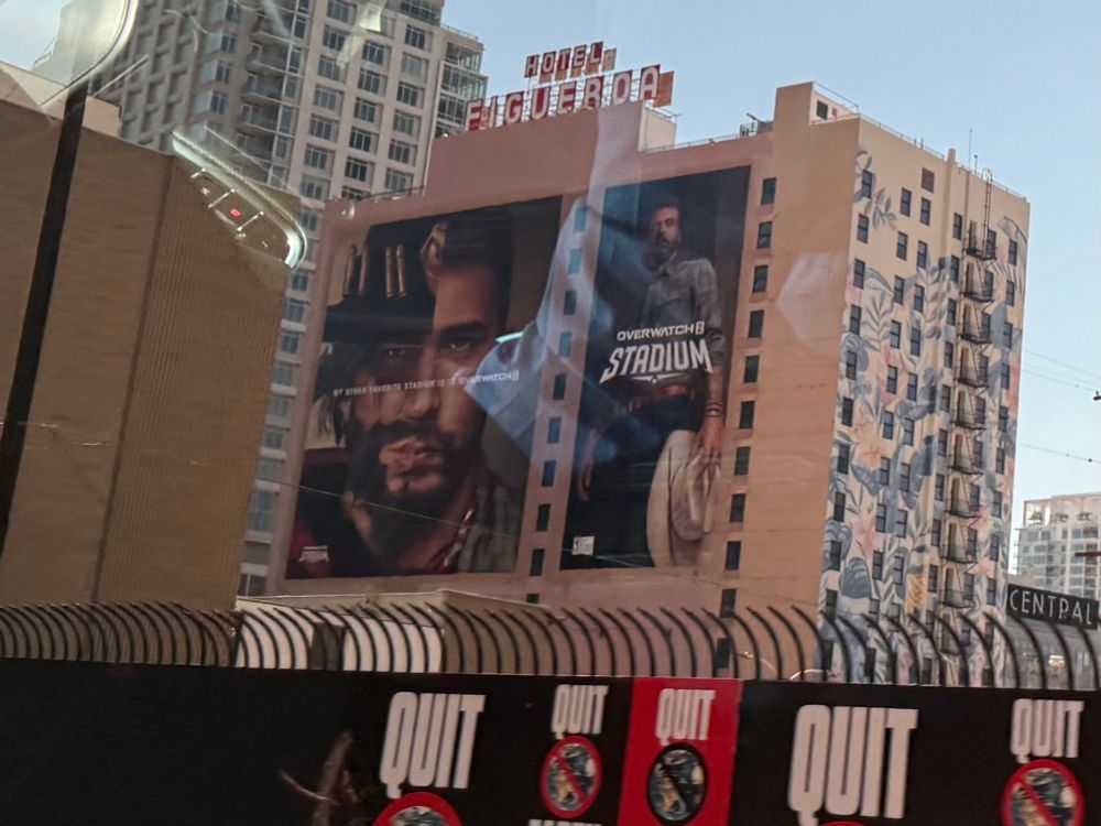 Photo of a brown building that is roughly 13 stories tall. The entire facade of one side of the building is covered by an advertisement for Overwatch 2