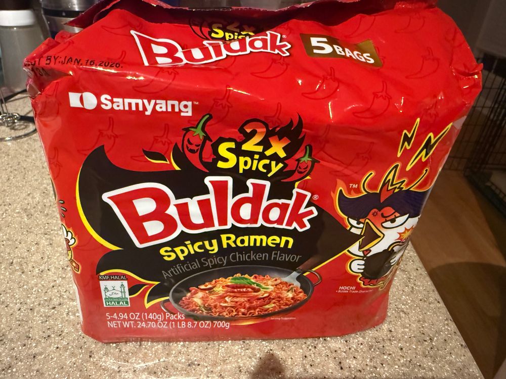 Photo of a 5-pack bag of Buldak brand 2x Spicy ramen. The package is red, with a chicken-like creature on the right looking very angry and breathing a black fire. Two chili peppers with evil smiles flank the words "2x Spicy." An image of deep red ramen in a black wok is visible at the bottom.