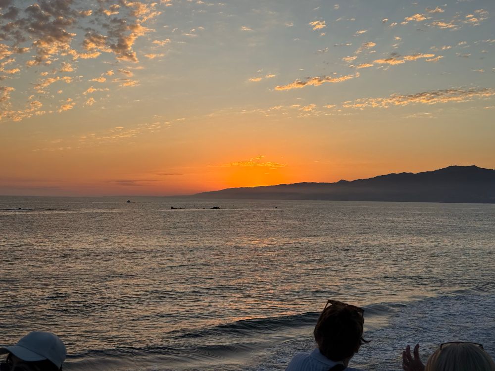 Photo of the Pacific Ocean from Santa Monica Pier, facing northwest toward the setting sun. The entire sun is just below the mountains visible in the background.