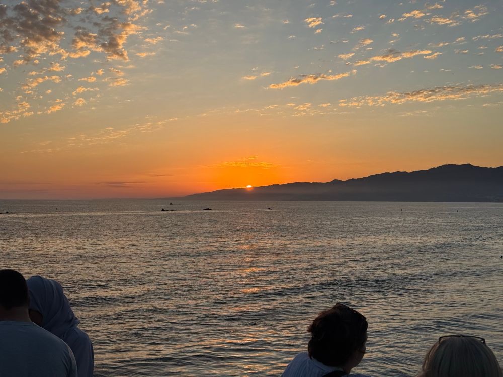 Photo of the Pacific Ocean from Santa Monica Pier, facing northwest toward the setting sun. Most of the sun is below mountains visible in the background, with only a sliver of the sun still visible.