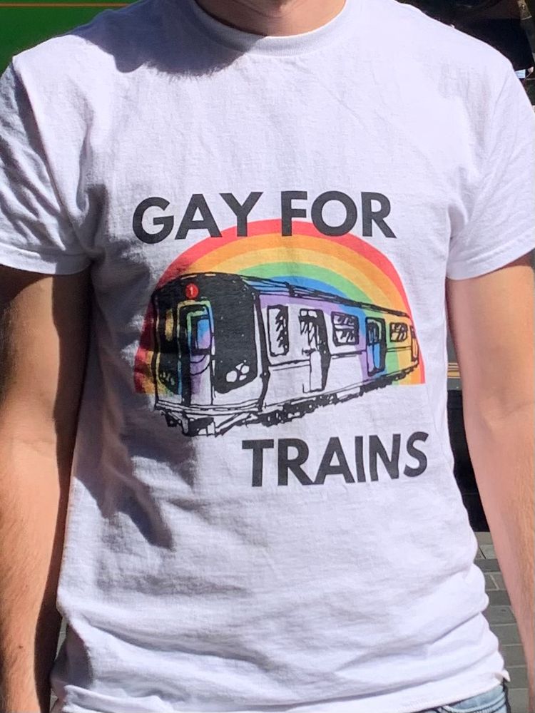 A close up of a T-shirt with a rainbow and a train carriage superimposed and the words ‘gay for trains’