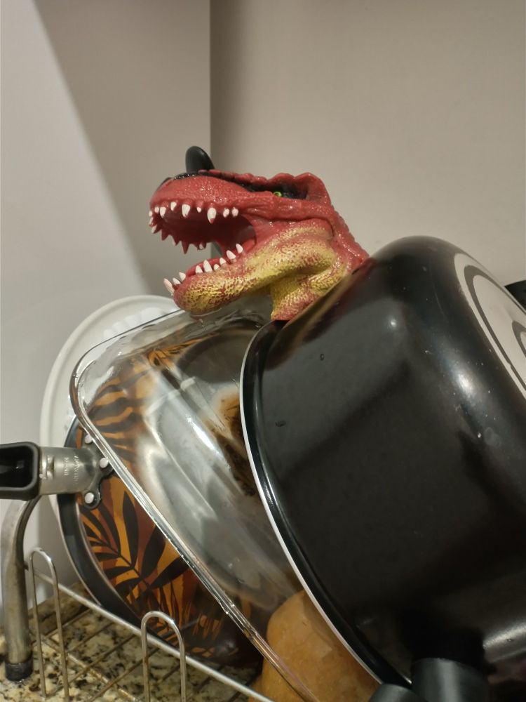 A imagem é fotografia de muitas louças no escorredor em cima da pia, e em cima delas está a cabeça de um fantoche de dinossauro