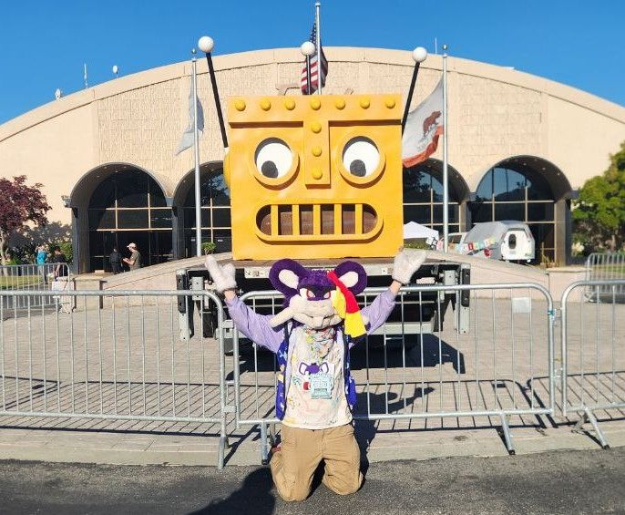 Photo of Rattata fursuit on their knees, arms raised, in front of the head of the robot mascot for Open Sauce.