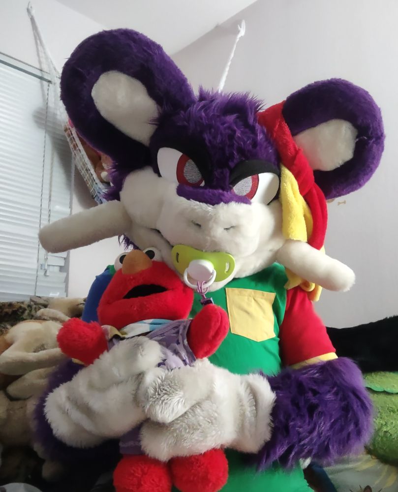 Photo of Rattata fursuit with a pacifier, holding a plush Elmo, in a colorblock onesie.