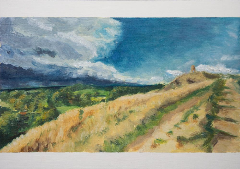 A painting of the top of Glastonbury Tor from a lower slope. The tower is in the distance on the left of the painting, atop a rise covered in brown grass. The slope winds its way along the bottom third of the painting, interspersed with green. On the left, the fields and woods in the distance are various shades of green. The top half of the painting is dominated by dark, stormy clouds on the left which lighten and give way to blue sunny skies on the right.