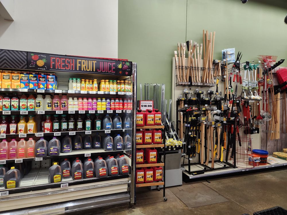 Photo of the back of my hometown grocery store, which shares space with an Ace Hardware. The shelves on the left have fresh fruit juice and sweet tea. The shelves on the right have gasoline tanks, hand axes, and other tools.