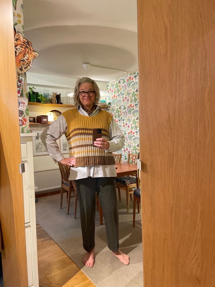 En helkroppsselfie i en spegel. Jag har en grov stickad randig väst över en ljus skjorta. Matplats i bakgrunden. 