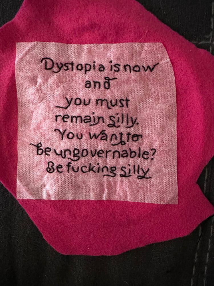 Hot pink fabric with black embroidered letters reading “dystopia is now and you must remain silly. You want to be ungovernable? Be fucking silly.”