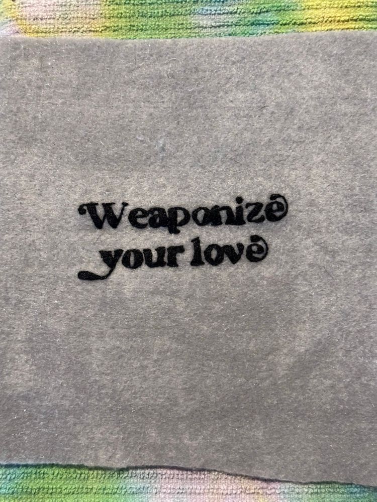 Finished embroidery, grey fabric and black thread. It reads “weaponize your love” 