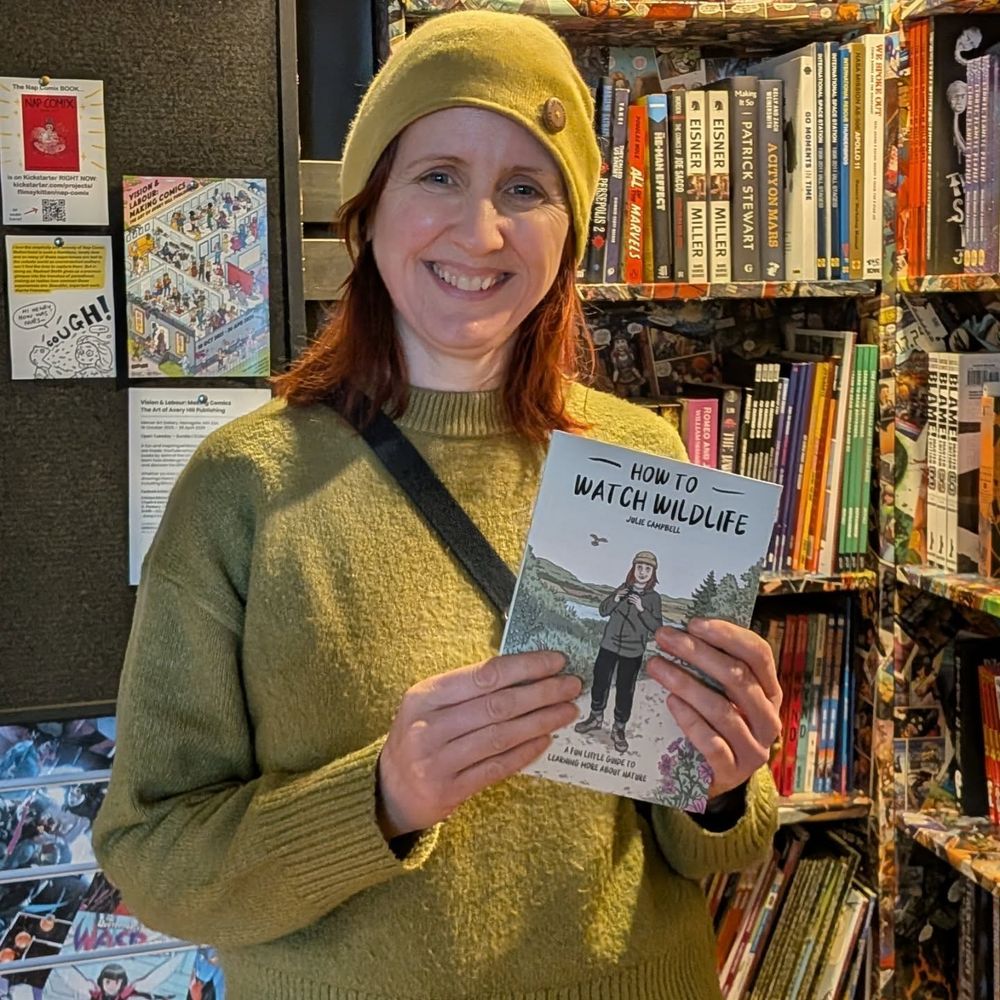 A smiling Julie Campbell stands in front of the shelves in Destination Venus holding a copy of her comic How to Watch Wildlife. 