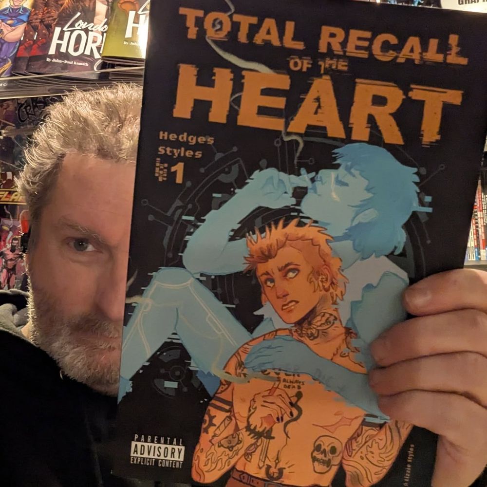 Regie holds a copy of Total Recall of the Heart to the camera. The cover has a black background featuring a tatooed shirtless young man, seen from the waist up in the foreground. He holds a lit cigarette in his right hand. Behind him is the blue apparition of a seated woman, also smoking. Her left arm is draped around his shoulder.
